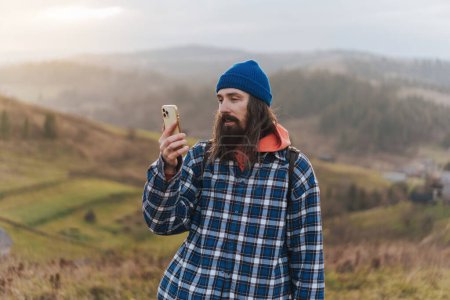 Photo for Traveler checking the map on smartphone while walking on hills wearing blue beanie, plaid shirt, orange sweatshirt and with a leather backpack on shoulders on sunset. Hiker male climber with a backpack behind his back stands against the backdrop of a - Royalty Free Image