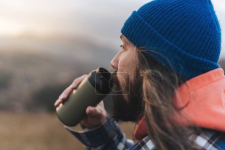 Photo for Portrait of photographer male drinking wather while making a stop during a walk the backdrop of a beautiful mountain landscape and looks into the distance.  Male traveler making a stop and enjoying the sunset while walking through the picturesque hil - Royalty Free Image