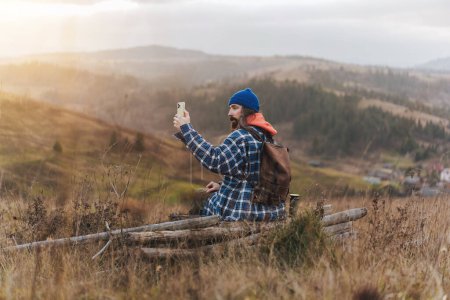 Foto de Vista desde la parte posterior de un joven fotógrafo de viaje barbudo con chaqueta azul tomando fotos en un teléfono inteligente mientras está sentado frente a verdes colinas borrosas durante el trekking en el campo de otoño al atardecer. - Imagen libre de derechos