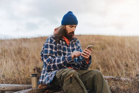 Photo for Young male travel photographer wearing blue beanie and jacket checking map on smartphone while a halt on the mountains on sunset. - Royalty Free Image