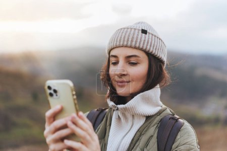 Téléchargez les photos : Portrait de voyageuse souriante heureuse utilisant un téléphone portable avec carte en ligne et textos à des amis, forêt floue et montagnes en arrière-plan, concept d'utilisation des technologies dans la nature - en image libre de droit