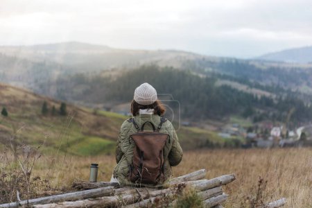 Foto de Vista desde la parte posterior de la chica excursionista con mochila y gorro de gorro sentado en el borde del acantilado y disfrutando de las montañas en la puesta de sol - Imagen libre de derechos