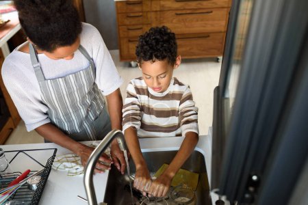 Téléchargez les photos : Fils aide mère dans la cuisine, laver les plats. Famille afro-américaine. - en image libre de droit