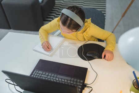 Photo for Cute child with headphones using laptop computer for online learning. Home school. Girl doing homework at home. Lifestyle concept for homeschooling. Social distancing and education. - Royalty Free Image