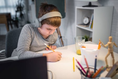 Foto de Lindo chico con auriculares haciendo la tarea en casa. Concepto de estilo de vida para la educación en el hogar - Imagen libre de derechos