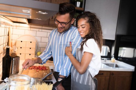 Foto de Feliz pareja multiétnica cocinando tomates caseros y pasta de albahaca. Cocinar en casa - Imagen libre de derechos