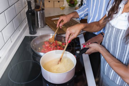 Foto de Feliz pareja multiétnica cocinando tomates caseros y pasta de albahaca. Cocinar en casa - Imagen libre de derechos