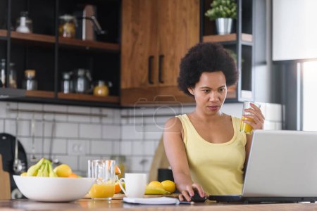 Téléchargez les photos : Jeune femme gaie surfer sur Internet sur un ordinateur portable à la maison dans la cuisine, et rafraîchissant avec une boisson de jus de fruits - en image libre de droit