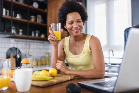 Foto de Joven mujer alegre navegando por Internet en una computadora portátil en casa en la cocina, y refrescante con una bebida de jugo de frutas - Imagen libre de derechos