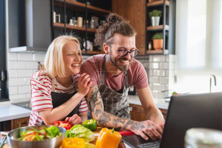Foto de Joven pareja alegre usando el teléfono inteligente mientras prepara un almuerzo vegano saludable en casa con computadora portátil - Imagen libre de derechos