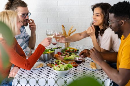 Photo for Young cheerful diverse people having lunch together at home. Toasting with Wine. Group of happy friends toasting while eating at the dining table. - Royalty Free Image