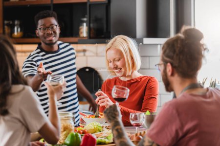 Téléchargez les photos : Jeunes gens diversifiés faisant une fête de vacances à la maison, préparant la nourriture, buvant, parlant et appréciant - en image libre de droit