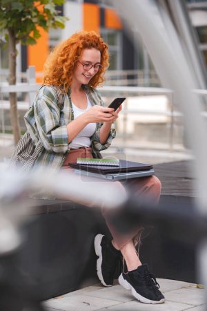 Téléchargez les photos : Belle étudiante rousse travaillant sur un ordinateur portable assis devant un bâtiment universitaire - en image libre de droit