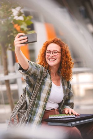Téléchargez les photos : Belle étudiante souriante aux cheveux roux prenant autoportrait avec un smartphone devant un bâtiment universitaire - en image libre de droit