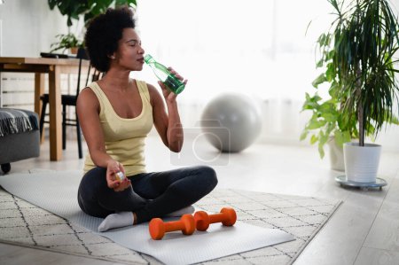 Téléchargez les photos : Femme afro-américaine faisant des exercices sur le sol à sa maison, prendre une pause et boire de l'eau. Hydratation. - en image libre de droit