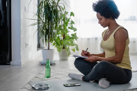 Photo for Young African-American woman writing in her notebook. Sitting at home by the window on a floor after exercise. Personal growth resolutions - Royalty Free Image