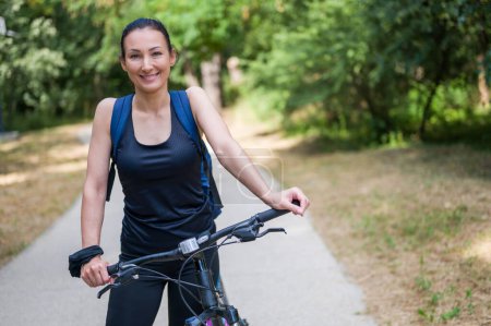 Téléchargez les photos : Portrait d'une cycliste souriante debout dans le parc à côté du vélo - en image libre de droit