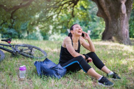 Téléchargez les photos : Femme souriante cycliste dans le parc public à l'aide d'un smartphone - en image libre de droit
