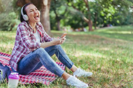 Téléchargez les photos : Femme avec écouteurs assis sur une herbe dans un parc public écoutant de la musique sur smartphone chantant fort. Émotions positives - en image libre de droit