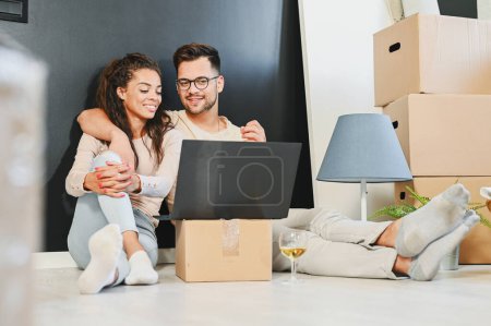 Photo for Happy multiethnic couple just moved into new empty apartment unpacking with laptop - Royalty Free Image