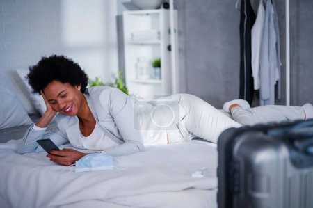 Téléchargez les photos : Jeune femme d'affaires noire vient d'arriver à l'hôtel pour se reposer d'un voyage, à l'aide d'un smartphone - en image libre de droit
