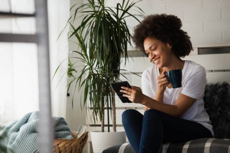 Téléchargez les photos : Jeune femme afro-américaine assise sur un canapé confortable au salon et utilisant une tablette numérique - en image libre de droit