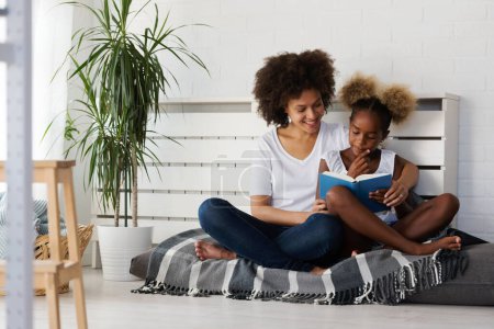 Téléchargez les photos : Belle mère africaine et fille lisant un livre ensemble à la maison. Coin confortable. - en image libre de droit