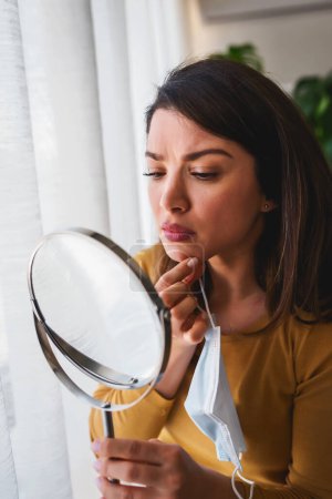 Photo for Woman looking herself in the mirror at home standing by the big window. She is concerned about acne - Royalty Free Image