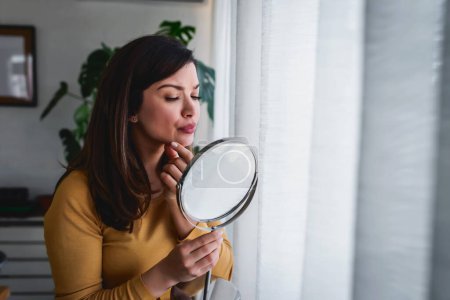 Photo for Woman looking herself in the mirror at home standing by the big window. She is concerned about acne - Royalty Free Image