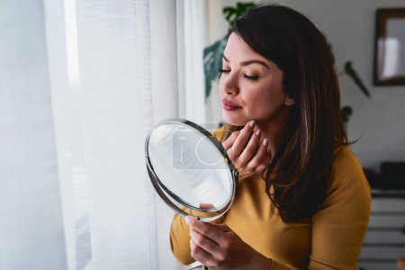 Téléchargez les photos : Femme se regardant dans le miroir à la maison debout près de la grande fenêtre. Elle est préoccupée par l'acné - en image libre de droit