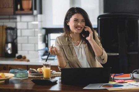 Téléchargez les photos : Jeune femme d'affaires souriante travaillant de la maison assise devant un ordinateur portable à la cuisine de la maison - en image libre de droit