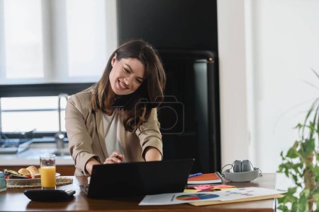 Photo for Young smiling businesswoman working from home sitting in front of laptop computer at home kitchen - Royalty Free Image