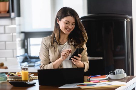 Téléchargez les photos : Jeune femme d'affaires souriante travaillant de la maison assise devant un ordinateur portable à la cuisine de la maison - en image libre de droit