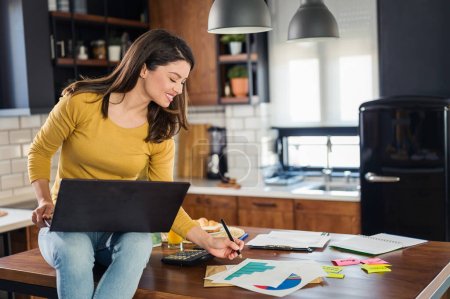 Téléchargez les photos : Jeune femme d'affaires souriante travaillant de la maison, assise à la maison cuisine avec ordinateur sur son ordinateur portable - en image libre de droit