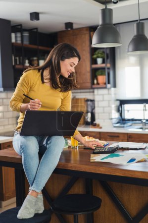Foto de Joven mujer de negocios sonriente trabajando desde casa, sentada en la cocina del hogar con computadora en su computadora portátil - Imagen libre de derechos
