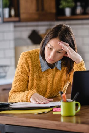 Téléchargez les photos : Femme d'affaires inquiète travaillant en ligne de la maison devant un ordinateur portable regardant dans les journaux et l'ordinateur. Stress - en image libre de droit