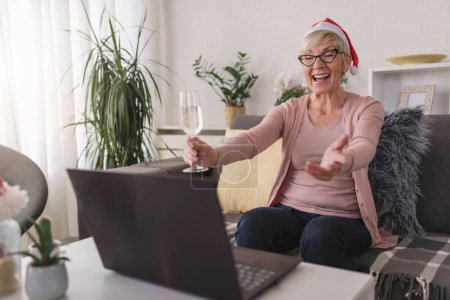 Photo for An old lady wearing a Santa hat drinking champagne alone at home toasting towards a computer. Making video calls over the internet with family during holidays. - Royalty Free Image