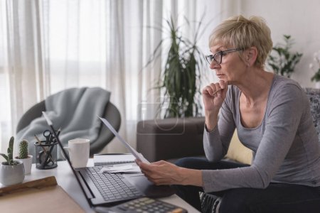 Ältere Rentnerin schaut sich Papiere an, berechnet Hypotheken, Ersparnisse und Ausgaben und plant ihr Eigenheimbudget