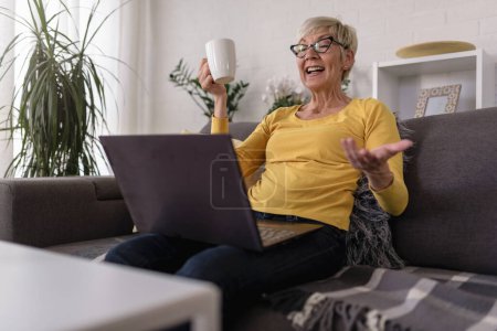 Photo for Beautiful senior elderly woman at home using laptop computer to communicate with her family, surfing internet, shopping online, drinking coffee - Royalty Free Image