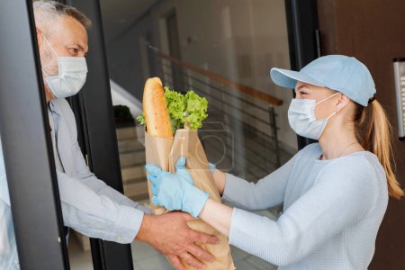 Foto de La chica del servicio de entrega trae comestibles a un hombre mayor durante la pandemia de COVID. peole usando máscaras protectoras. - Imagen libre de derechos