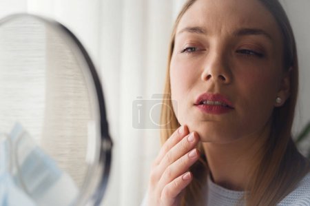 Téléchargez les photos : Jeune belle femme se regardant dans le miroir à la maison. Inquiète pour l'acné. Problèmes d'acné. - en image libre de droit