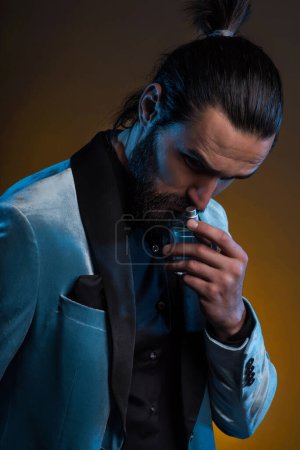 Foto de Hermoso hombre joven barbudo masculino sosteniendo una botella de fragancia. Perfume masculino, hombre barbudo en traje. - Imagen libre de derechos
