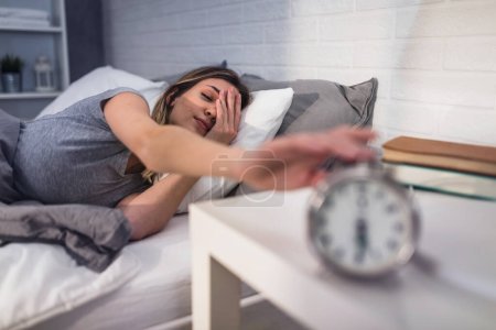Téléchargez les photos : Une belle jeune femme se réveille tôt le matin dans sa maison ensoleillée en cherchant un réveil - en image libre de droit
