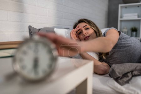 Photo for A beautiful young woman is waking up early in the morning in her sunny home reaching for an alarm clock - Royalty Free Image