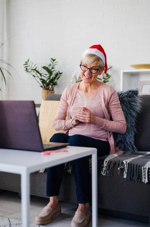 Photo for An old lady wearing a Santa hat drinking champagne alone at home toasting towards a computer. Making video calls over the internet with family during holidays. - Royalty Free Image