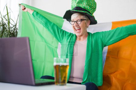 Foto de Vieja irlandesa mirando el partido, bebiendo cerveza, animando a Irlanda con una bandera irlandesa y un sombrero de duende. Ganar un juego! - Imagen libre de derechos