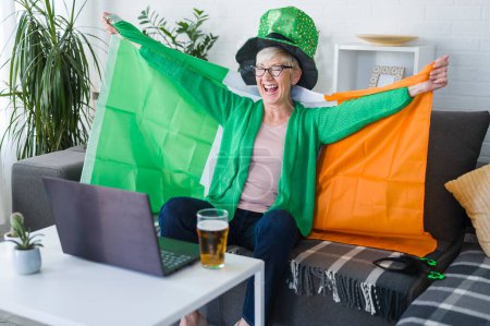 Téléchargez les photos : Vieille Irlandaise regardant le match, buvant de la bière, acclamant l'Irlande avec un drapeau irlandais et un chapeau de lutin. Gagner un match! - en image libre de droit
