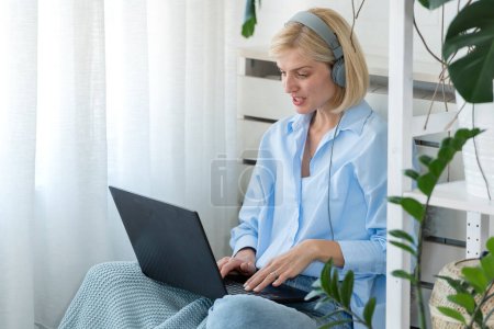 Téléchargez les photos : Jeune femme ayant des cours de langue en ligne à la maison à l'aide d'un ordinateur portable et d'écouteurs, assis sur un sol près d'une grande fenêtre. Éducation à domicile en ligne - en image libre de droit
