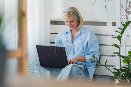 Téléchargez les photos : Jeune femme ayant des cours de langue en ligne à la maison à l'aide d'un ordinateur portable et d'écouteurs, assis sur un sol près d'une grande fenêtre. Éducation à domicile en ligne - en image libre de droit