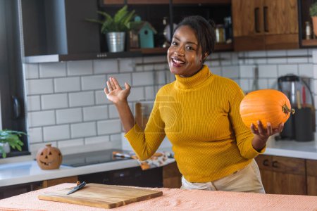 Photo for Young smiling black woman preparing for Halloween carving a pumpkin at home in the kitchen - Royalty Free Image